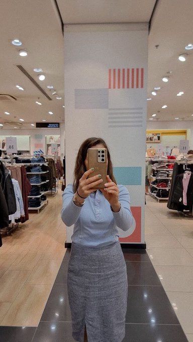 A mirror selfie of a woman in a clothing store wearing a light blue button-up top and a gray long skirt, holding a phone to take the photo.