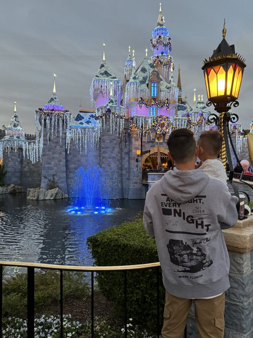A person wearing a grey 'An Every Night, A No Cover Night, Main Florida' hoodie holds a small child while they both look at an illuminated, icicle-draped castle at a theme park.
