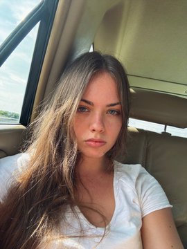 A young woman with long brown hair and blue eyes sits inside a car, taking a selfie while wearing a white t-shirt.