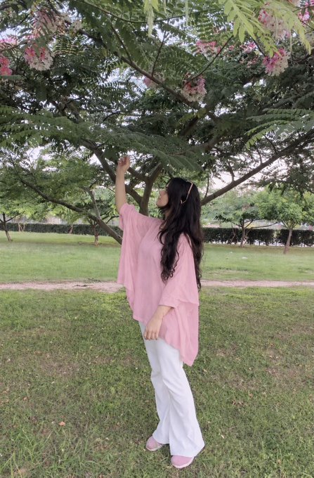 A woman in a pink poncho and white pants standing in a park, reaching up toward pink flowers on a tree.