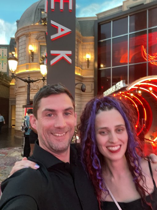 A man and a woman with purple-streaked dreadlocks smile for a selfie in front of the 'IMAX' theater and 'STREAMING STARS' neon signage.