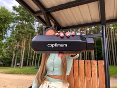A person wears a black and pink cat-ear 'Optimum' VR headset under a wooden shelter with trees in the background.