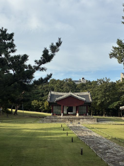 A traditional Korean pavilion with a green tiled roof and red accents sits at the end of a stone path on a manicured lawn surrounded by pine trees.