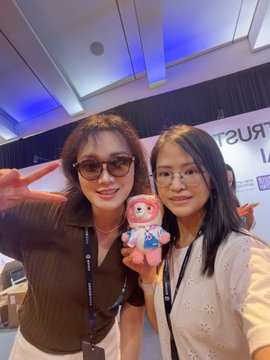 Two women pose together indoors, with one wearing sunglasses and making a peace sign, while holding a small plush mascot toy between them.