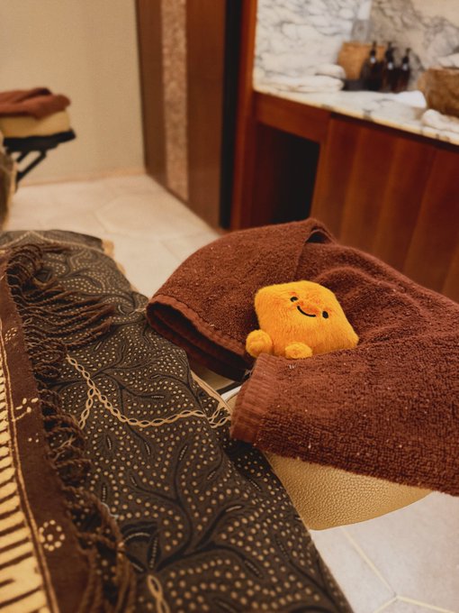 A small yellow smiley-face plush toy rests on a folded brown towel beside a patterned brown blanket, set on what appears to be a massage table in a spa-like room.