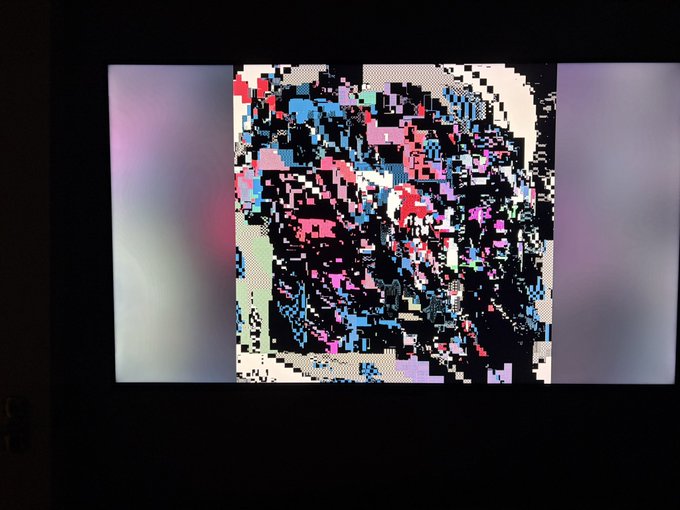 A screen displaying heavily pixelated, glitchy artwork in vivid pink, blue, and black colors against a dark background.