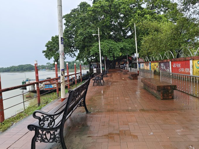 The image shows a park near a river with several benches placed along its walkway, offering visitors a place to sit and enjoy the view of the water. The benches are made of metal and have an old-fashioned design. In addition to the benches, there is also a boat visible in the background on the left side of the image. A post can be seen at the top right corner of the photo, which appears to contain some text or information related to the park's location or features.