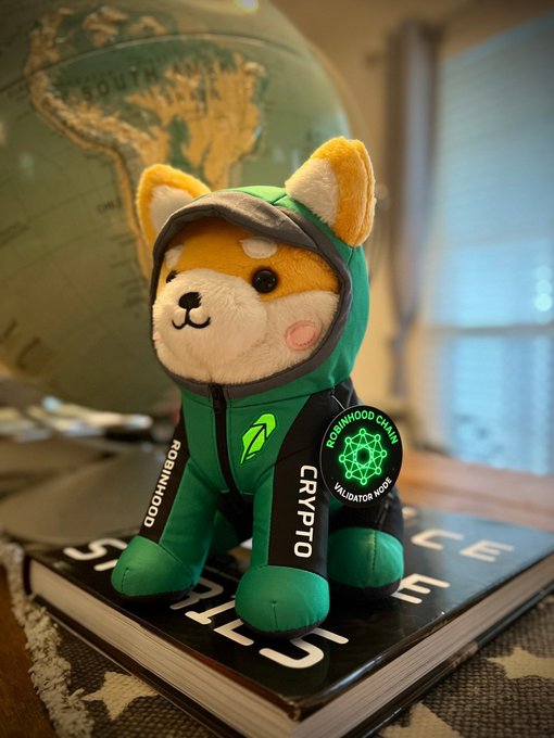 A plush Shiba Inu toy dressed in a green "Crypto" hoodie and a "Robinhood Chain Validator Node" tag sits on a book next to a globe, visually echoing the post's mention of Robinhood Chain and the $ASTEROID token contract address.