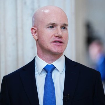 A close-up portrait of a bald man wearing a dark suit, white shirt, and a blue tie, with a small black microphone clipped to his lapel.