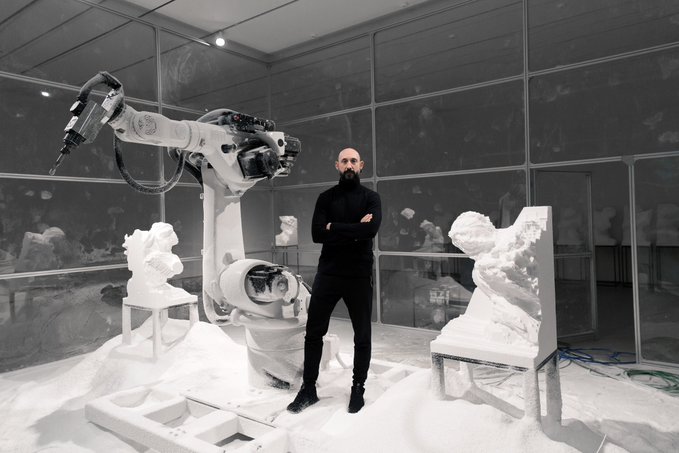 The image features a man standing next to a white robot that appears to be an art installation. The man is dressed in black and has a beard, giving him a more mature or artistic appearance. The room he's standing in seems to be a workshop filled with art equipment. There are two chairs in the scene – one near the man on the left side of the image and another closer to the right. The background shows an assembly line-like structure that looks like it could be part of the installation itself. In addition, there is a sculpture visible at the far end of the room, located behind the man who is standing.