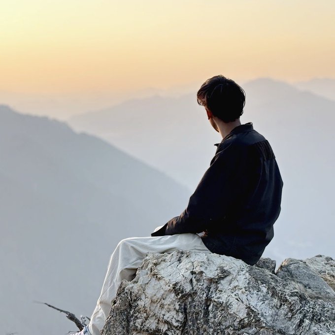 A young man with dark hair sits on a rocky outcrop, gazing at a misty mountain landscape during what appears to be sunrise or sunset.
