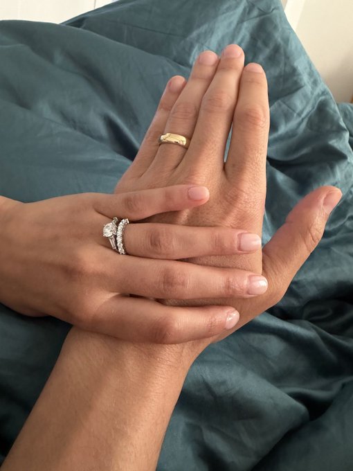 A close-up photo of two hands clasped together on a teal blanket, both wearing diamond rings, suggesting an engagement or commitment.