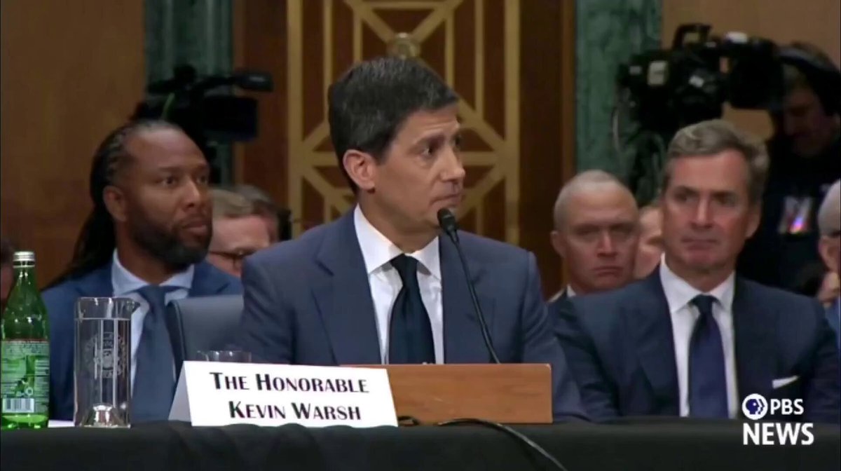 The image shows a man giving a speech at a podium, with a TV screen behind him displaying the news coverage of his announcement. The man speaking has a name tag that reads "KEVIN WARSH", indicating he might be an important figure or a guest speaker. The TV screen also displays the words "NEW FED CHAIR KEVIN WARSH ANNOUNCED NOT TO CREATE A CENTRAL BANK DIGITAL CURRENCY". This news coverage suggests that Kevin Warsh has made a significant announcement related to digital currency, which could be an important development in the world of finance and technology.

The image also includes several other people present at the event, some sitting on chairs while others stand around the podium. There are multiple TVs mounted on walls behind the speakers, likely displaying live footage or images from the event. A few bottles can be seen placed nearby, possibly containing water for the attendees to stay hydrated during the speech.
