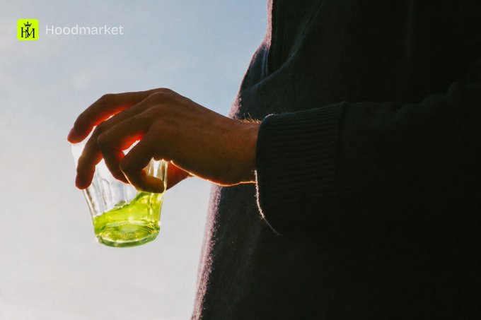 A silhouetted hand holds a small glass of green liquid against a bright sky, with the "Hoodmarket" logo visible in the corner, evoking the mystery of unlocking NFT assets. The image's cryptic, almost ceremonial vibe mirrors the post's tease about getting closer to revealing one's digital holdings.