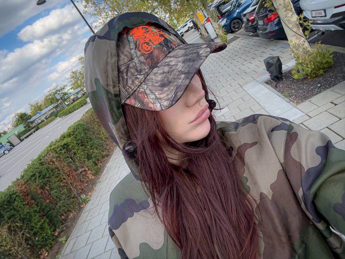 A young woman with long dark hair takes a selfie outdoors while wearing a camouflage hoodie and a cap featuring an orange Harley-Davidson logo, with a parking lot visible in the background.