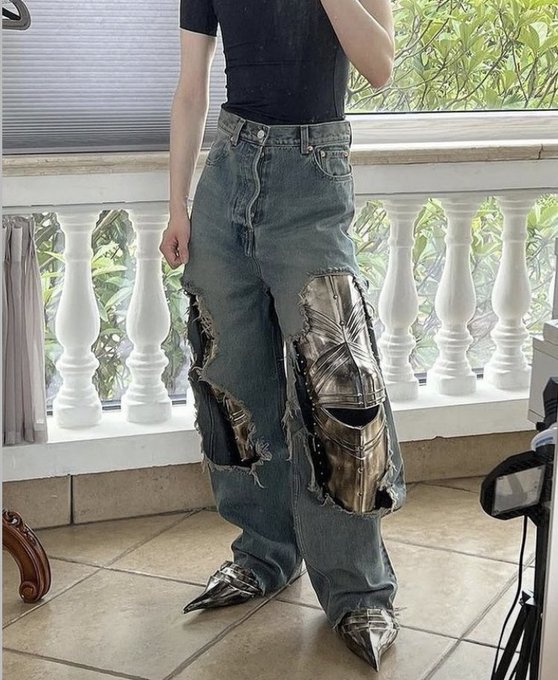 A person models heavily distressed denim jeans with large metallic silver patches and matching pointed metallic silver boots.