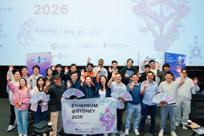A group photo of event attendees holding fans and a banner that reads 'ETHEREUM @SYDNEY 2026, Sydney · Sep 26 - 27', with a large screen behind them displaying the event title and sponsor logos.