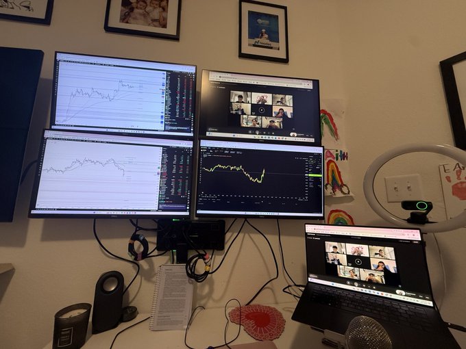 A home trading setup featuring four monitors displaying stock charts and a video conference call, alongside a laptop showing the same video call, with a ring light and rainbow decorations nearby.