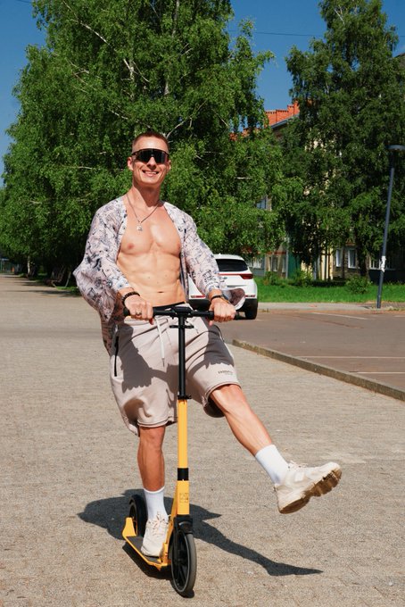A muscular shirtless man wearing sunglasses, a floral shirt, and beige shorts posing on a yellow scooter on a sunny street with trees and a white SUV in the background.
