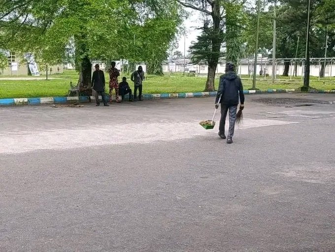 A photograph showing a person walking across a paved area carrying a basin and a broom, while others sit in the background near some trees.