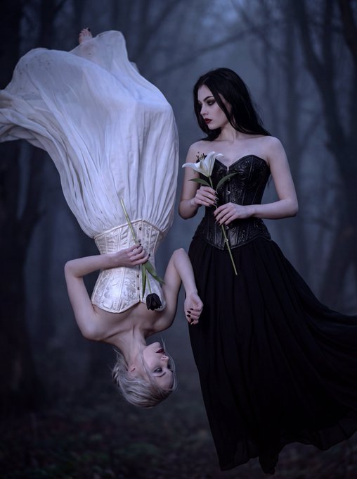 Two women pose in a misty forest setting: one stands upright in a black gothic gown holding a white lily, while the other hangs upside down in a flowing white dress holding a wilting flower.