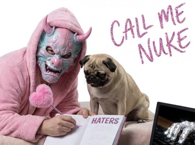 A promotional-style image featuring a person in a pink hooded robe and pink devil horn mask writing in a notebook labeled "HATERS" with a heart-shaped pen, accompanied by a pug, with the text "CALL ME NUKE" and a laptop showing a gloved hand.
