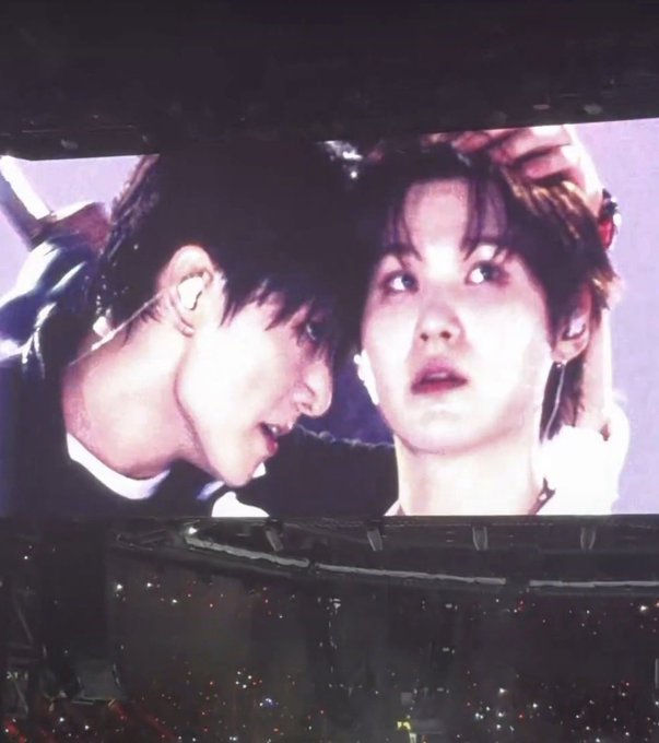 A photo taken at a concert showing two male performers close together on a large screen above a darkened audience with lights.