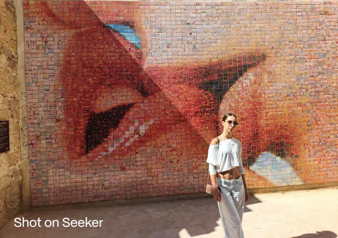 A woman in a light blue two-piece outfit and sunglasses poses in front of a large red mosaic mural, with 'Shot on Seeker' watermark at the bottom left.