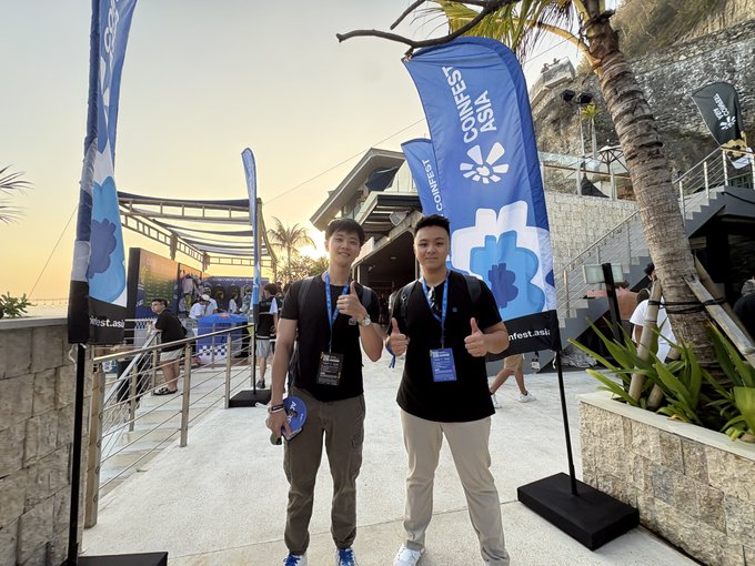 Two young men giving thumbs up at an outdoor event with 'LEAPFEST ASIA' blue flags, palm trees, and a sunset sky in the background, with a venue and other attendees visible.