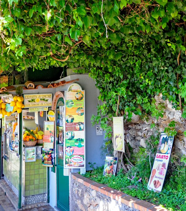 The image shows a small outdoor market with various fruits, including oranges and bananas, displayed for sale. The market has a green door and a stone wall surrounding it. There are several signs posted outside of the market, possibly advertising the products available or providing information about the market's offerings.