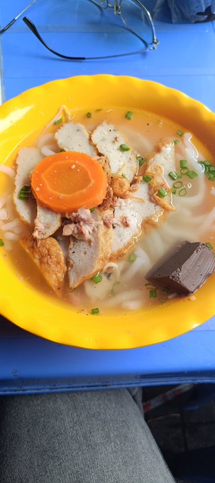 A yellow bowl filled with noodle soup containing sliced meat, a carrot slice, meatball pieces, tofu, and green onions, sitting on a blue table.