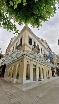 A street-level view of a corner building with a yellow facade and an ornate white metal awning over the sidewalk, partially shaded by green tree branches overhead.