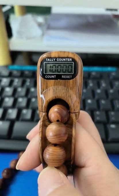 A hand holds a wooden digital tally counter displaying '00000' with 'COUNT' and 'RESET' buttons, attached to wooden prayer beads, with a black computer keyboard in the background.