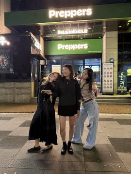 Three members of the BNB Chain MKT Team pose together outside "Preppers" in Seoul, smiling warmly as they promote their upcoming October 2nd event, with the brightly lit storefront signage visible behind them.