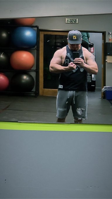 Mirror selfie of a muscular man in a gym wearing a black tank top, gray ripped jeans, and a cap, taking a photo with his phone.