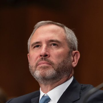 A middle-aged man with gray hair and a beard, wearing a dark suit and blue tie, is seated in a formal setting with a wood-paneled background.
