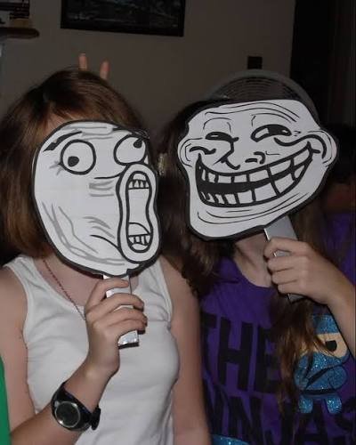 Two girls holding up printed Trollface meme masks on sticks, with one expressing shock and the other grinning mischievously.
