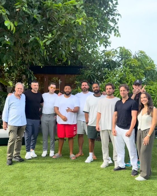 A group of nine people stands together on a grassy lawn in front of a building surrounded by trees, posing for a group photo in casual summer attire.