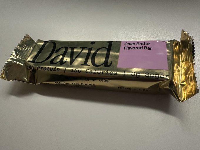 A gold foil-wrapped David protein bar labeled 'Cake Batter Flavored Bar' sits on a light surface, showing '28g Protein | 150 Calories | 0g Sugar'.