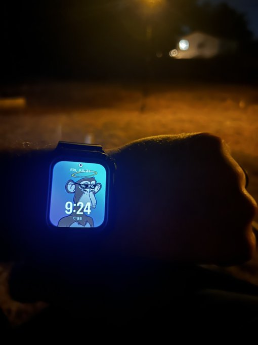 A smartwatch on a wrist displays a cartoon monkey face (resembling a Bored Ape) with the time "9:64," date "FRI, JUL 31," and temperature "86°," set against a dim outdoor backdrop with distant lights—visually reinforcing the message of staying connected despite life's busyness.