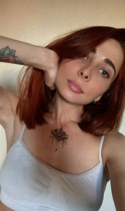 A young woman with red hair poses in a selfie, wearing a light blue crop top, with a floral tattoo on her arm and a geometric rose tattoo on her chest.