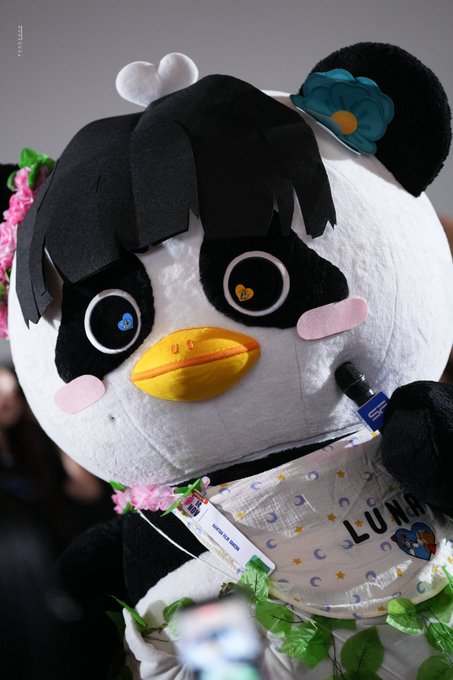 A person in a large penguin mascot costume with a flower on its head appears at an event, holding a small electronic cigarette device.