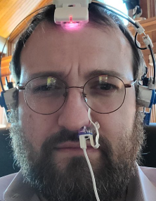 A close-up of a bearded man with glasses wearing a complex technological headset with lights and a facial sensor attachment.