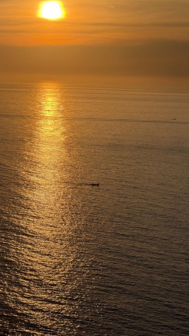 A serene sunset over the ocean with golden light reflecting on the water and a small boat visible in the distance.