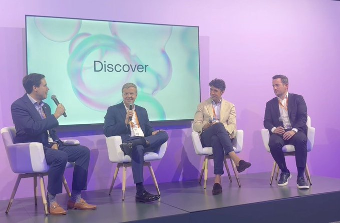 Four men sit on stage during a panel discussion with a 'Discover' display screen behind them.