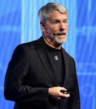 A photograph of Michael Saylor, the MicroStrategy founder known for Bitcoin advocacy, speaking at a presentation while wearing a black blazer over a dark shirt with an ear-mounted microphone.