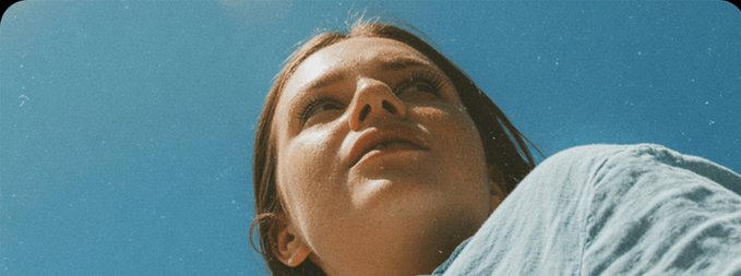 A low-angle portrait of a young woman with brown hair looking upward against a clear blue sky, wearing a light blue top.