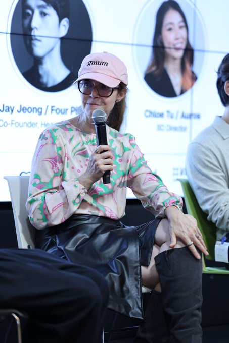 A woman wearing a pink 'muum' cap and floral blouse speaks into a microphone at a panel, with 'Jay Jeong / Four...' and 'Chloe Yu / Aur... BC.Zen...' visible on the screen behind her.