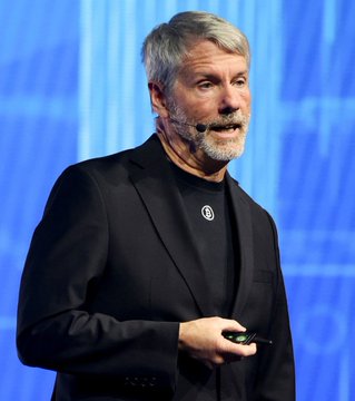 A gray-bearded man with a headset microphone, wearing a black blazer over a black shirt, speaks on stage while holding a presentation remote, likely referencing Tom Lee, the Fundstrat co-founder mentioned in the post about significant unrealized losses on crypto holdings.