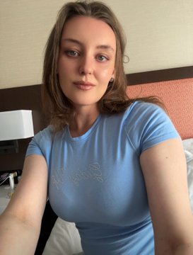 A woman with shoulder-length light brown hair wearing a light blue t-shirt takes a selfie in what appears to be a hotel room.